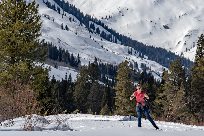 Photo of a woman nordic skiing on a groomed trail with snowy mountains in the background.