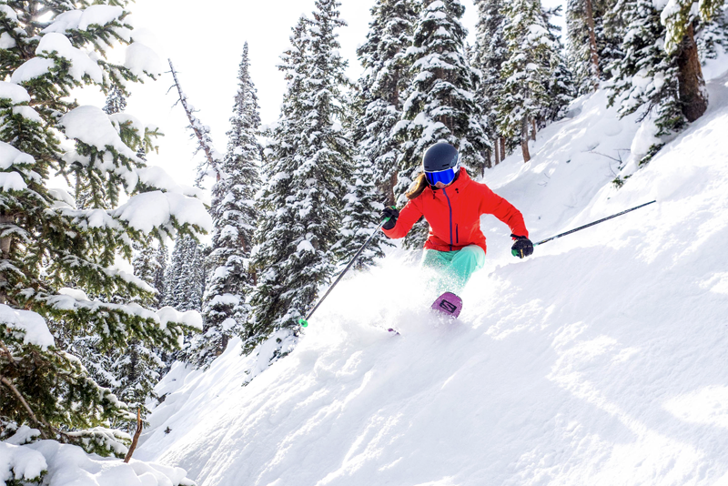 Photo of a woman in a red jacket skiing powder on steep terrain with trees.